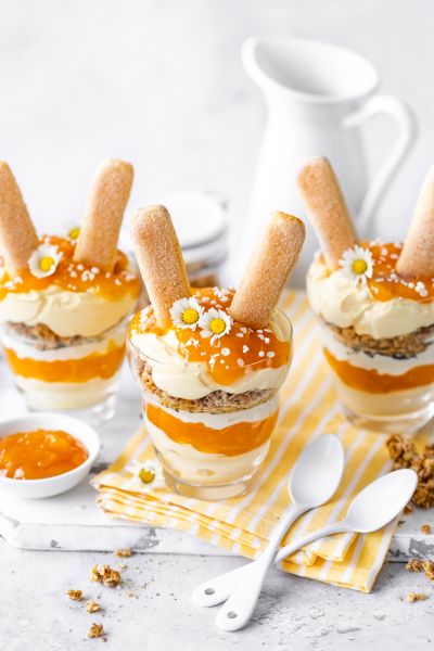 Schichtdessert mit Eierlikör, Maintal Bio Aprikose-Mango und Granola, serviert in Gläsern mit Biskiutstäbchen als Osterhasenohren. Serviervorschlag auf Kaffefetafel.