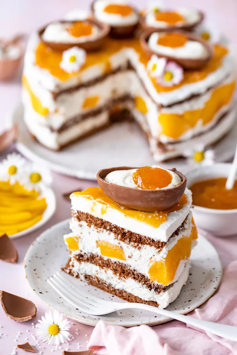 Ostertorte Schoko-Aprikose-Mango mit Löffeleiern Stück der Ostertorte Schoko-Aprikose-Mango mit Löffeleiern auf Teller, dahinter die Torte auf Platte.