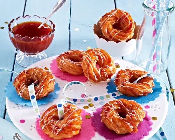 Spritzkringel mit Hagebuttenglasur Serviervorschlag Spritzkringel mit hagebuttenglasur auf Tortenplatte, Luftschlangen, Schüssel mit Hagebutten-Konfitüre mit Löffel und Glaskaraffe mit Röhrchen.
