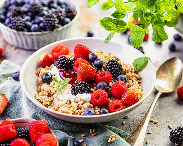 Frühstücksbowl mit Dinkel und Bio Gartenfrüchten Frühstücksbowl mit Dinkel und Bio Gartenfrüchten in Bowlschüssel mit Löffel, daneben zwei Schüsseln jeweils mit frischen Früchten.