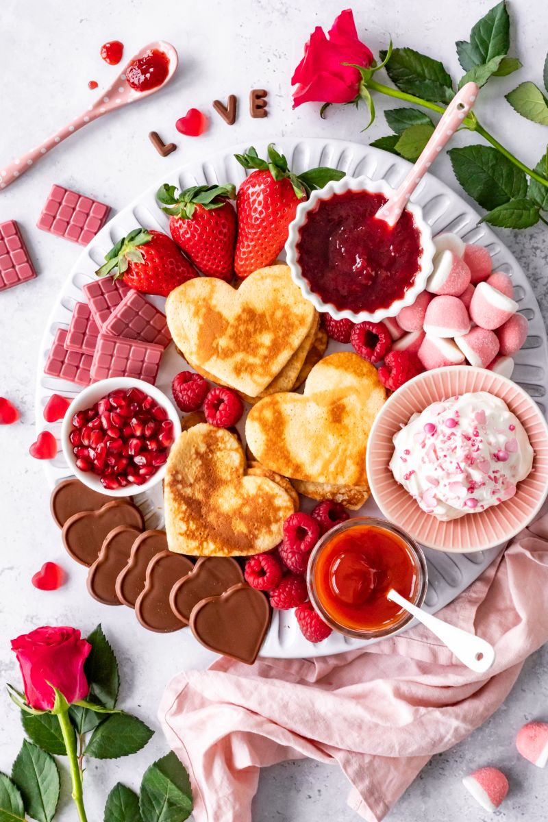 Pancake-Board mit Herz-Pfannkuchen auf Teller. Daneben Rosen und das Wort Love aus Schokoladenbuchstaben.