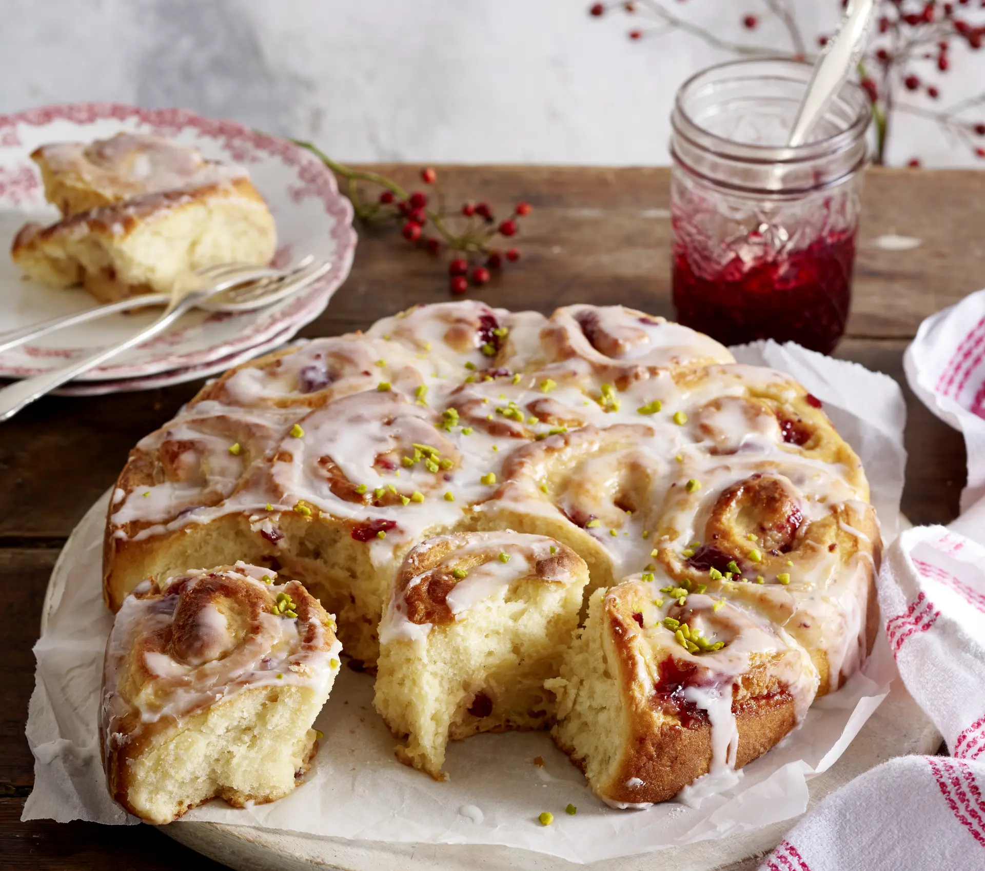 Cranberry-Pudding-Schneckenkuchen mit Zuckerguss und gehackten Pistazien, angeschnitten auf einer Holzplatte serviert.