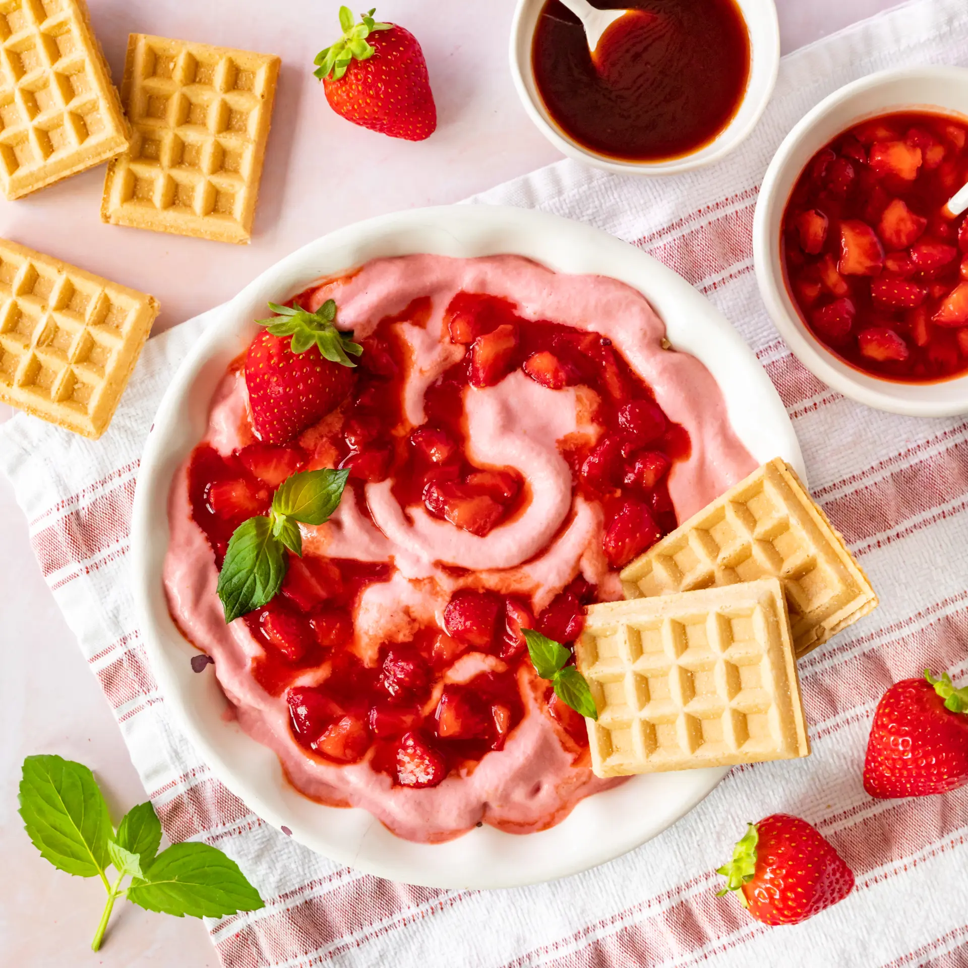 Cremiges rosa Hüttenkäse-Eis auf einem Teller, garniert mit Erdbeerstücken und Hagebutten-Soße, dekoriert mit frischen Erdbeeren, Minze und Waffeln.