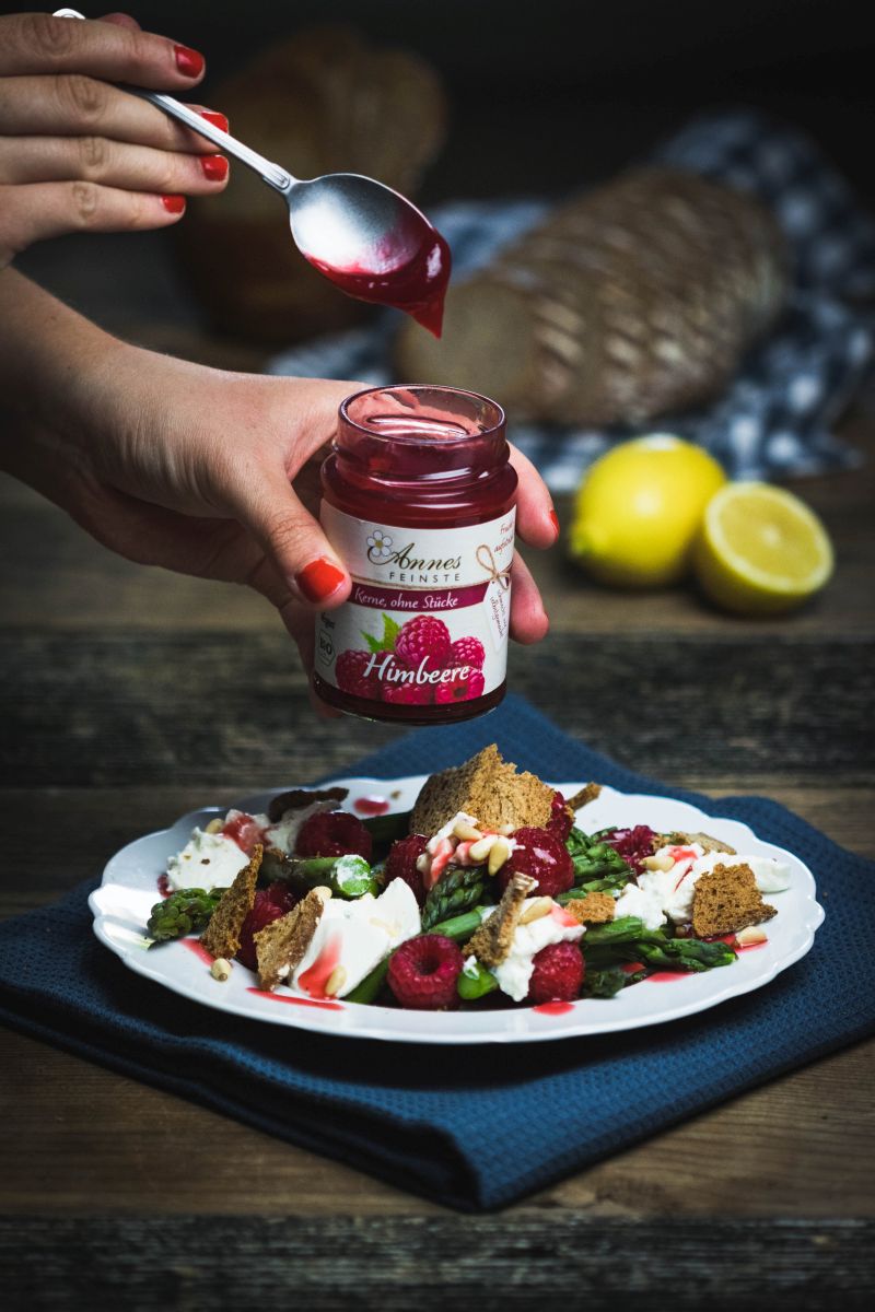 Himbeer-Spargelsalat mit Ricotta und Sauerteigbrotchips Annes Feinste Himbeer-Fruchtaufstrich wird mit Löffel auf Teller mit Himbeer-Spargel-Salat getropft.