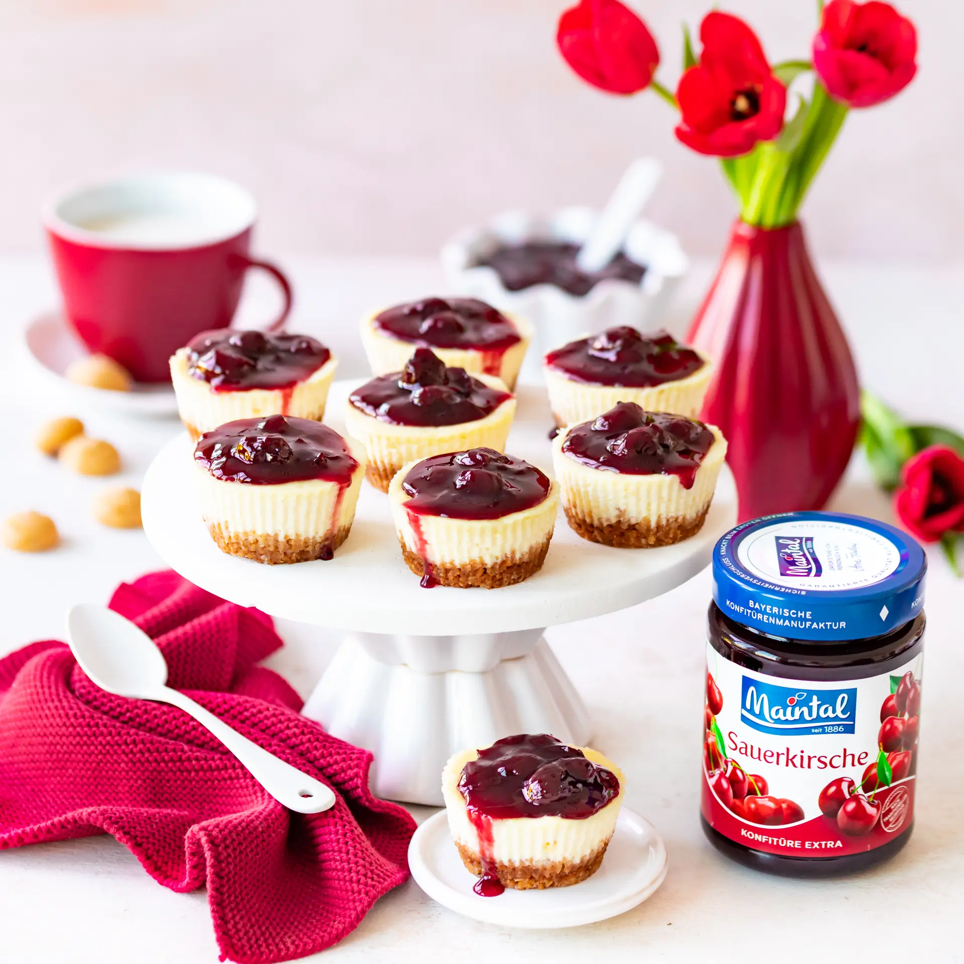 Mini-Cheescakes mit Kirschen Mehrere Mini-Cheesecakes mit Amaretti-Boden und Sauerkirsch-Konfitüre auf einer weißen Tortenplatte, daneben ein Glas Maintal Sauerkirsch-Konfitüre und rote Tulpen in einer Vase.