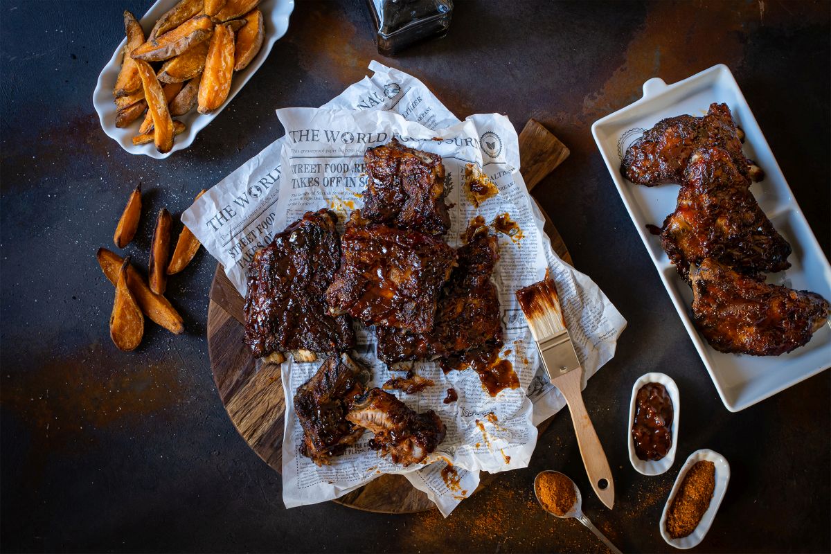 Serviervorschlag Spare-Ribs aus dem Ofen mit Hagebutten-Barbecuesauce auf Backpaperier und in der Schale mit Schale Pommes, Saucen und Currypulver.