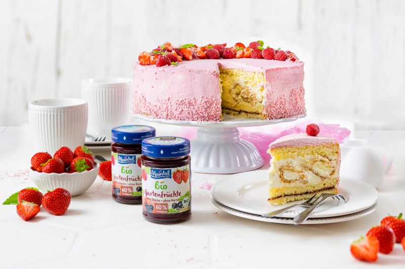 Windbeuteltorte mit Gartenfrüchten Tisch mit Windbeuteltorte mit Gartenfrüchten auf Tortenplatte, Teller mit einem Stück der Torte, Kaffeetassen, Schüssel mit frischen Erdbeeren und zwei Gläsern Maintal Bio Gartenfrüchte Fruchtaufstrich.