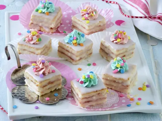 Faschings-Petit-Fours mit Hagebutten-Konfitüre, weißer Glasur und bunten Zuckerguss-Tuffs in Rosa und Blau.