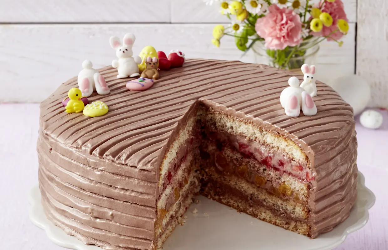 Ostertorte mit Schoko-Creme und Frucht Dekorierte Ostertorte mit Schokocreme, Frucht und Osterdeko auf Platte, dahinter Blumen.