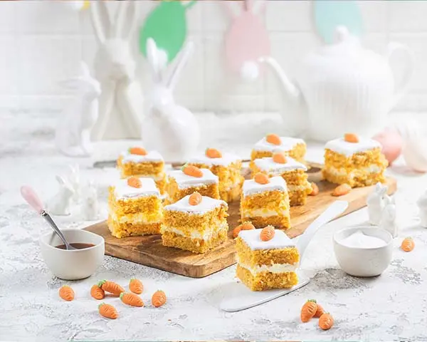 Mehrschichtige Rüblikuchenwürfel mit weißem Zuckerguss und dekorativen Marzipanmöhren, auf einem hellen Tisch angerichtet.