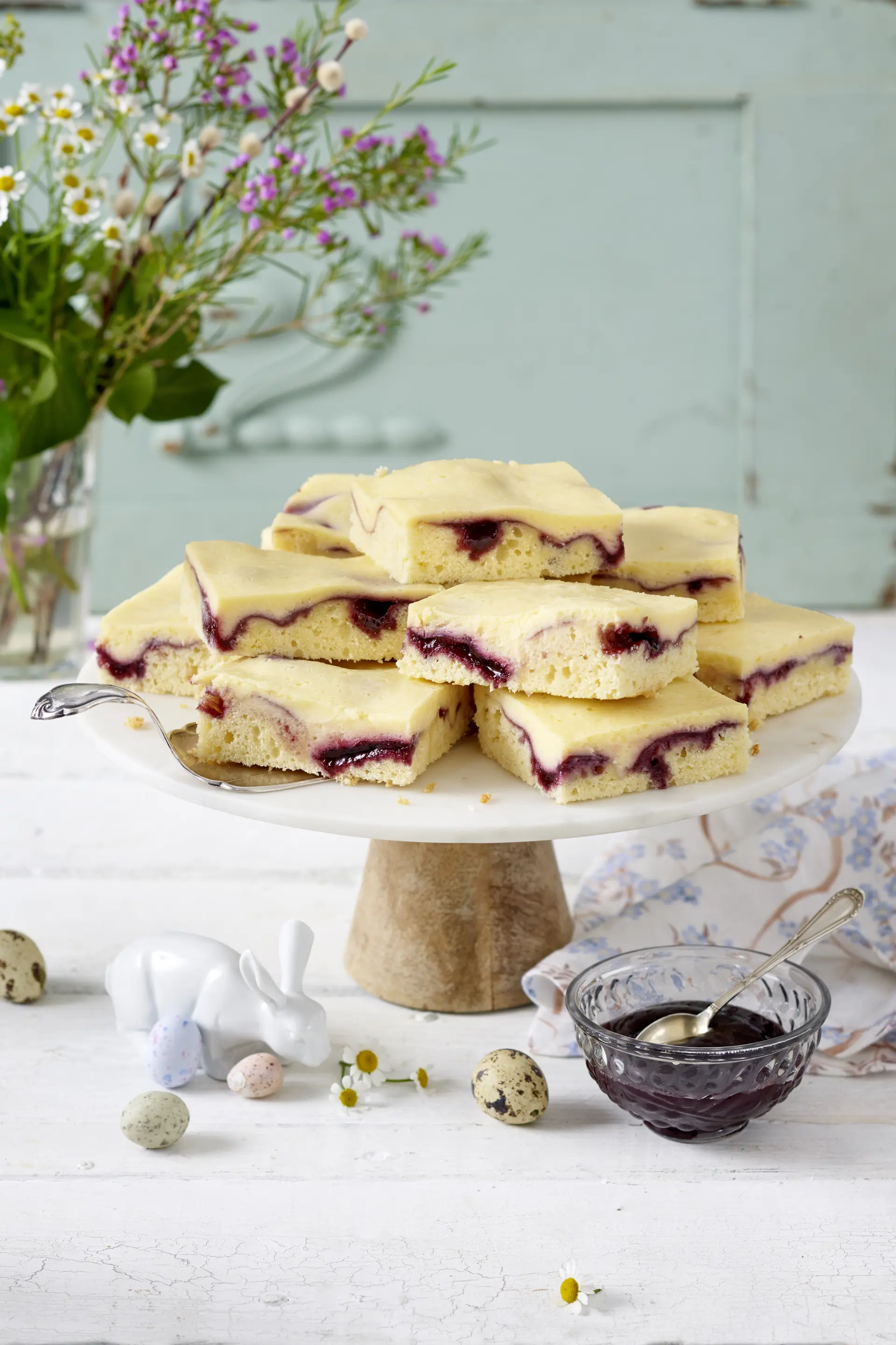 Fruchtige Buttermilch-Schnitten mit Waldfrucht Buttermilch-Schnitten mit Waldfruchtaufstrich auf einer Tortenplatte, frühlingshaft mit Blumen, Ostereiern und Deko-Hase arrangiert.