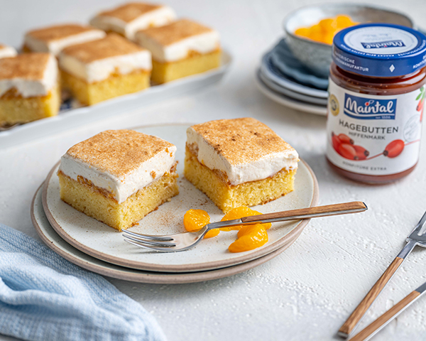 Blechkuchen mit Schmand-Sahne-Creme und Mandarinen Zwei Stücke Schmand-Sahne-Creme und Mandarinen auf Teller mit Gabel, daneben Glas Hagebutten-Konfitüre.