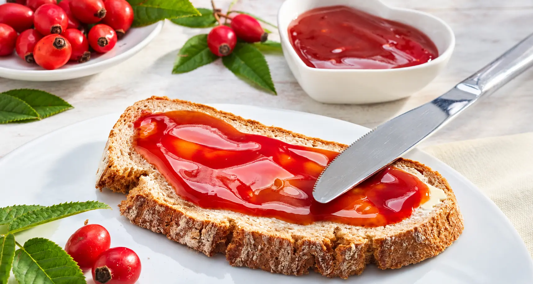 Scheibe Brot mit Butter und roter Hagebutten-Konfitüre auf einem Teller, daneben frische Hagebutten und eine Schale Konfitüre.