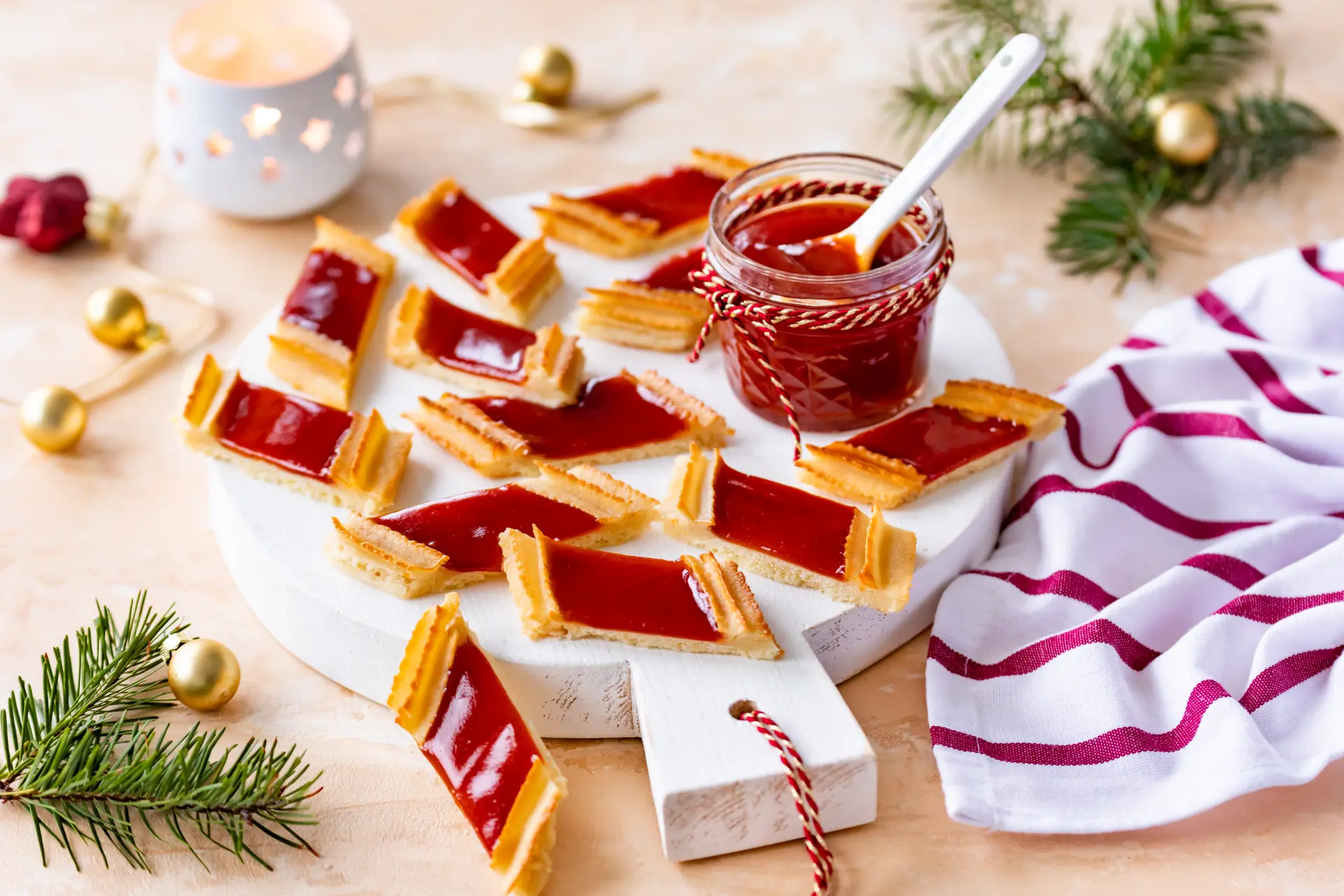 Marzipanplätzchen mit roter Hagebutten-Konfitüre auf einem Holzbrett, weihnachtlich dekoriert mit Tannenzweigen und Kugeln.