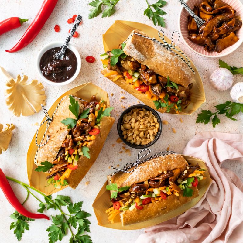 Banh Mi-Sandwich, vegan Draufsicht auf drei Banh-Mii-Sandwiches in Bambusschälchen serviert, Schälchen mit Pflaumenmus, Erdnüsse und marinierte Pilzen, Chilies und Koriander.