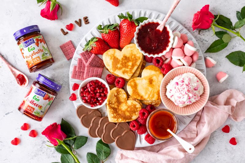 Serviervorschlag Pancake-Board mit Herz-Pfannkuchen auf Teller, daneben Maintal Bio Fruchtaufstriche und Rosen.