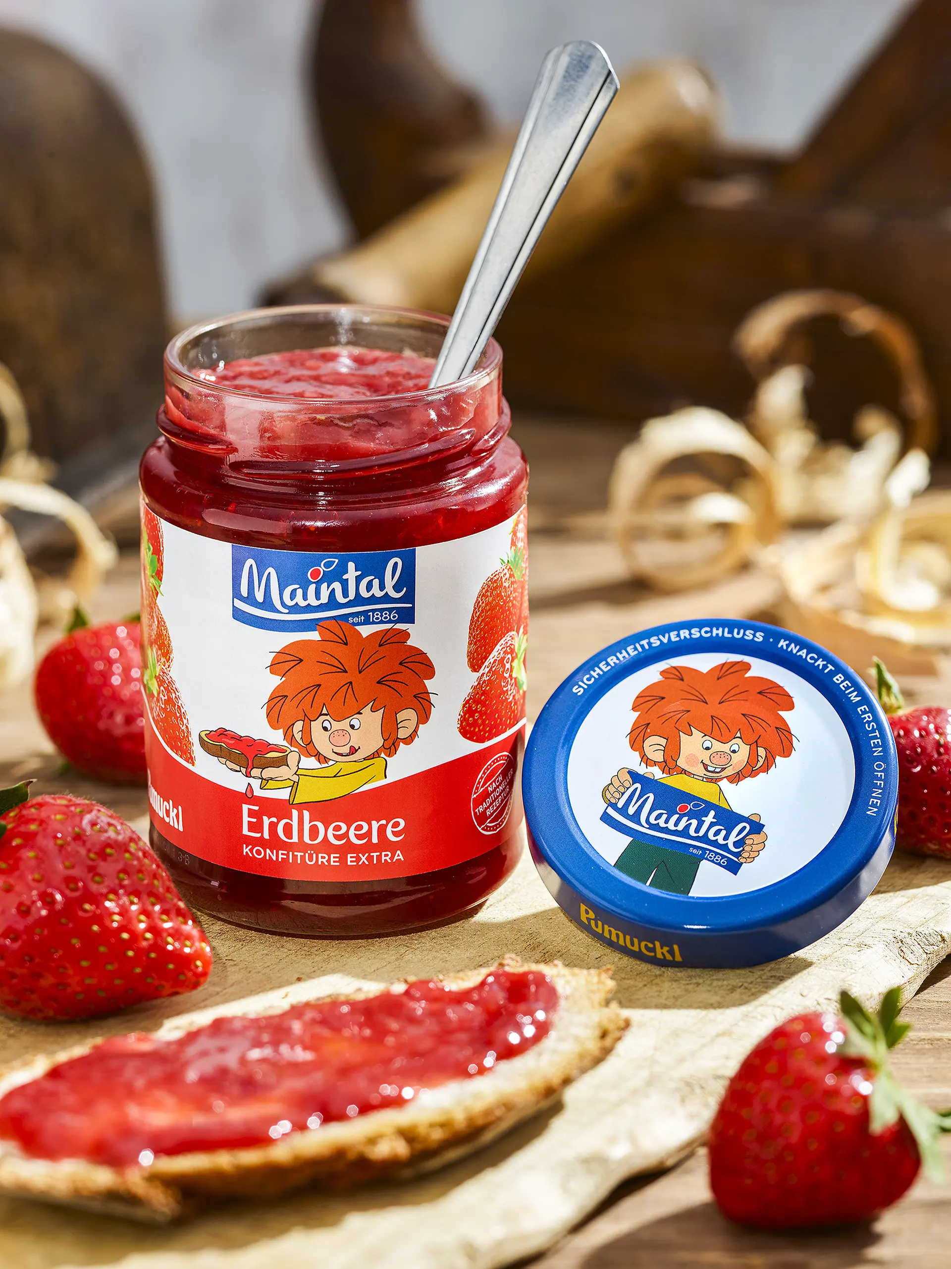 Erdbeer-Konfitüre extra Offenes Glas Erdbeerkonfitüre mit Löffel, Deckel daneben, Erdbeeren und Brot mit Konfitüre auf Holztisch.