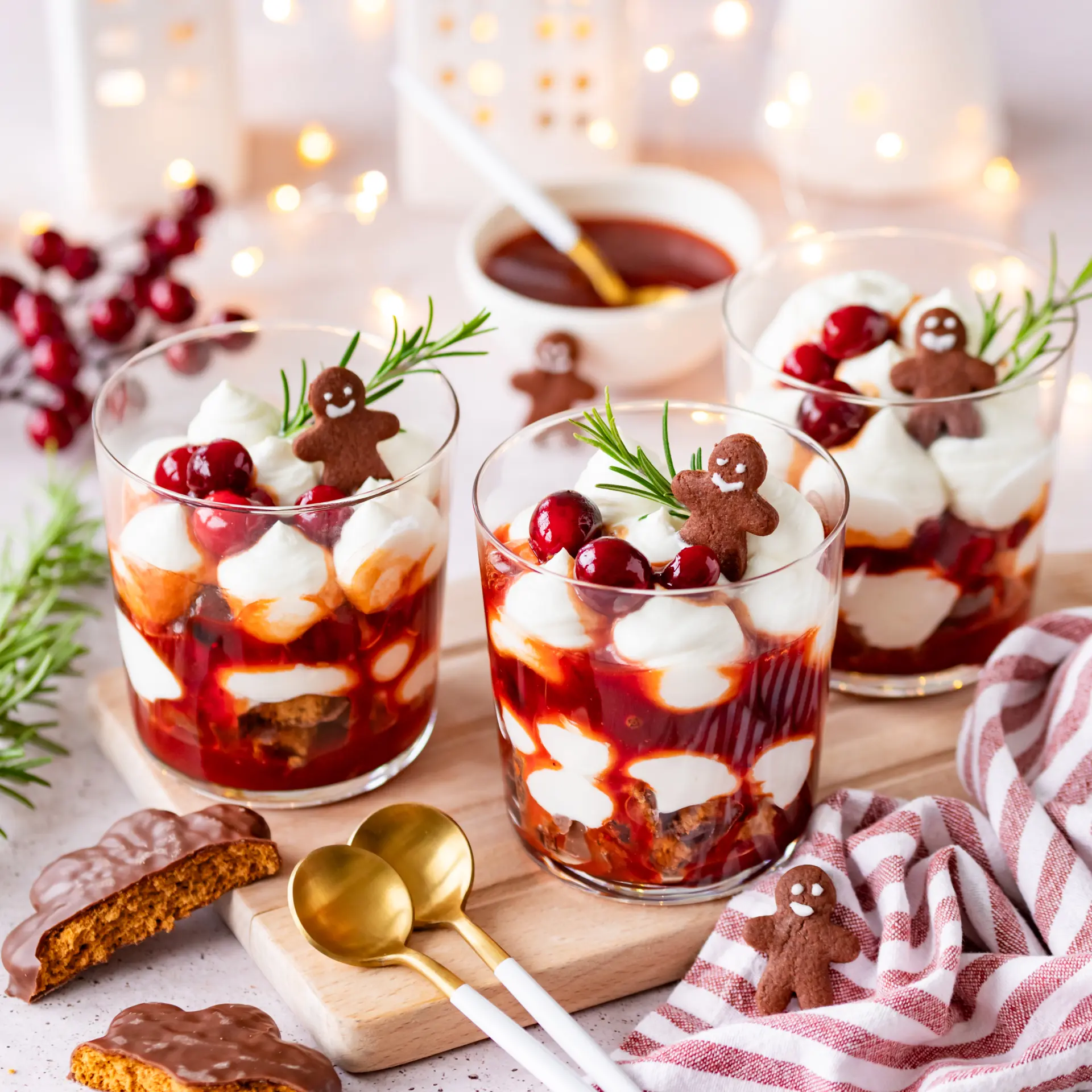 Drei Gläser mit geschichtetem Lebkuchen-Dessert, roter Fruchtsauce und weißer Creme. Oben Cranberries, Rosmarin und kleine Lebkuchenfiguren. Festliche Deko im Hintergrund.