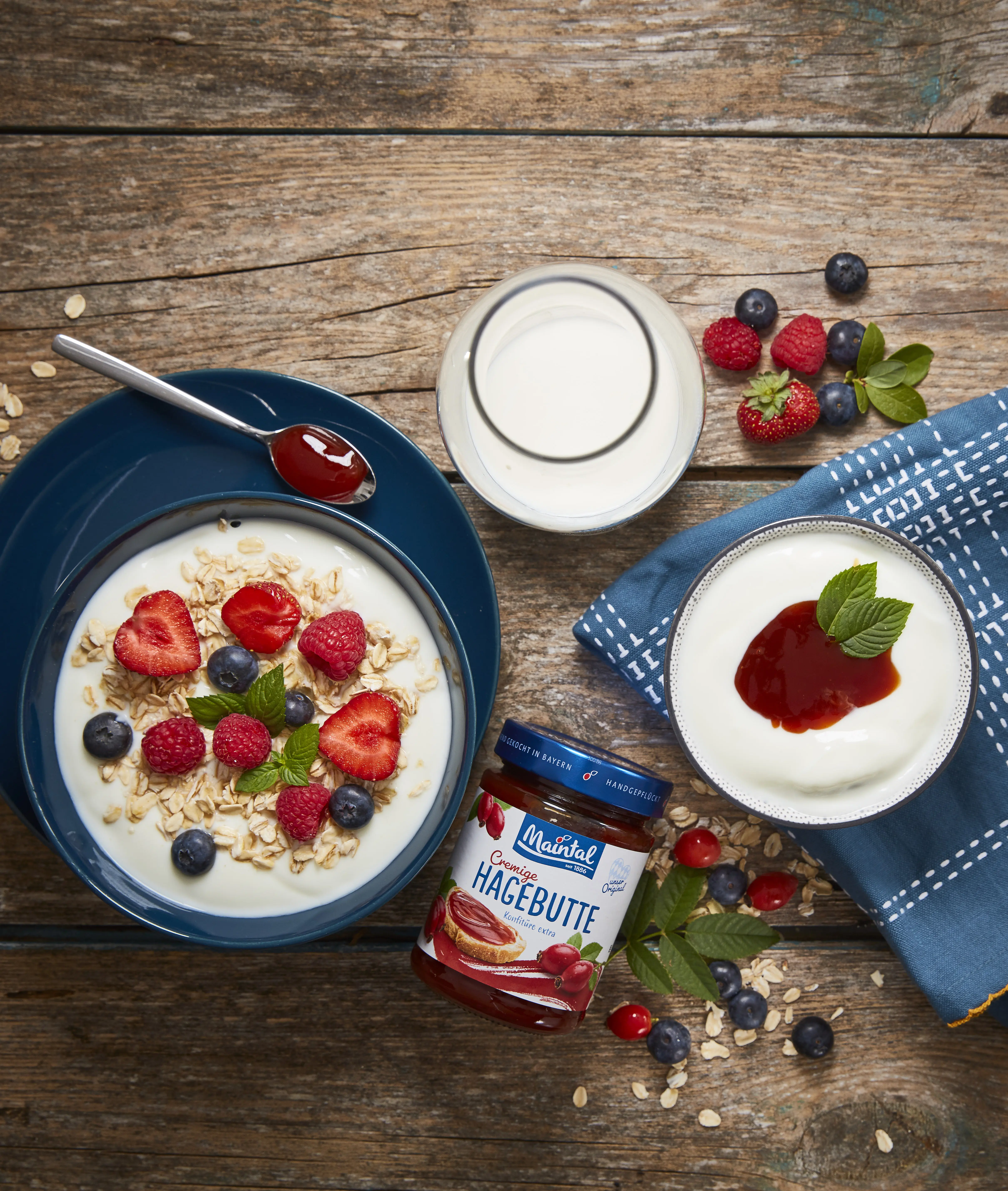 Joghurt mit Haferflocken, Beeren und Hagebutten-Konfitüre auf einem Holztisch, daneben Milch und ein Glas Hagebutten-Konfitüre.