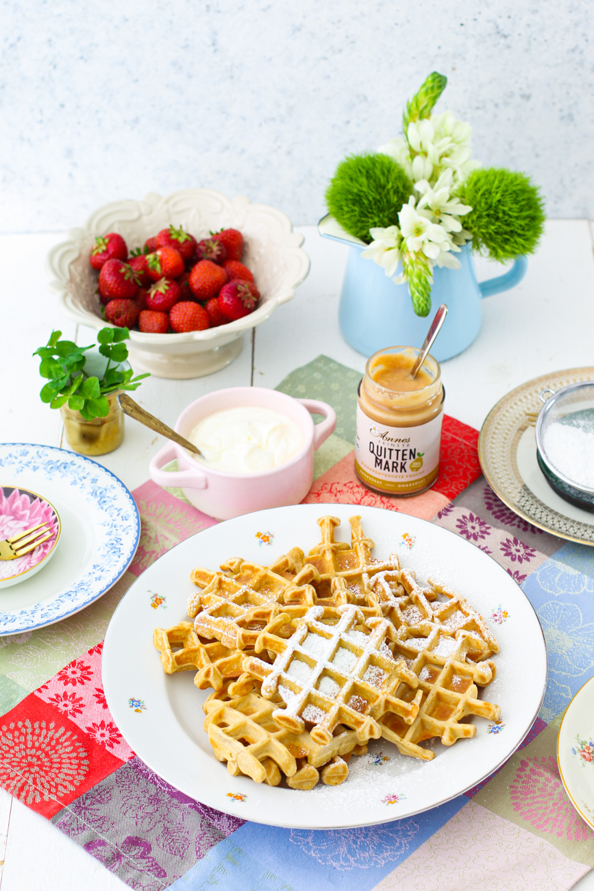 Teller mit Waffeln. Dahinter steht ein Glas Quittenmark mit Loeffel, eine Schuessel Puderzucker, frische Erdbeeren in einer Schuessel.