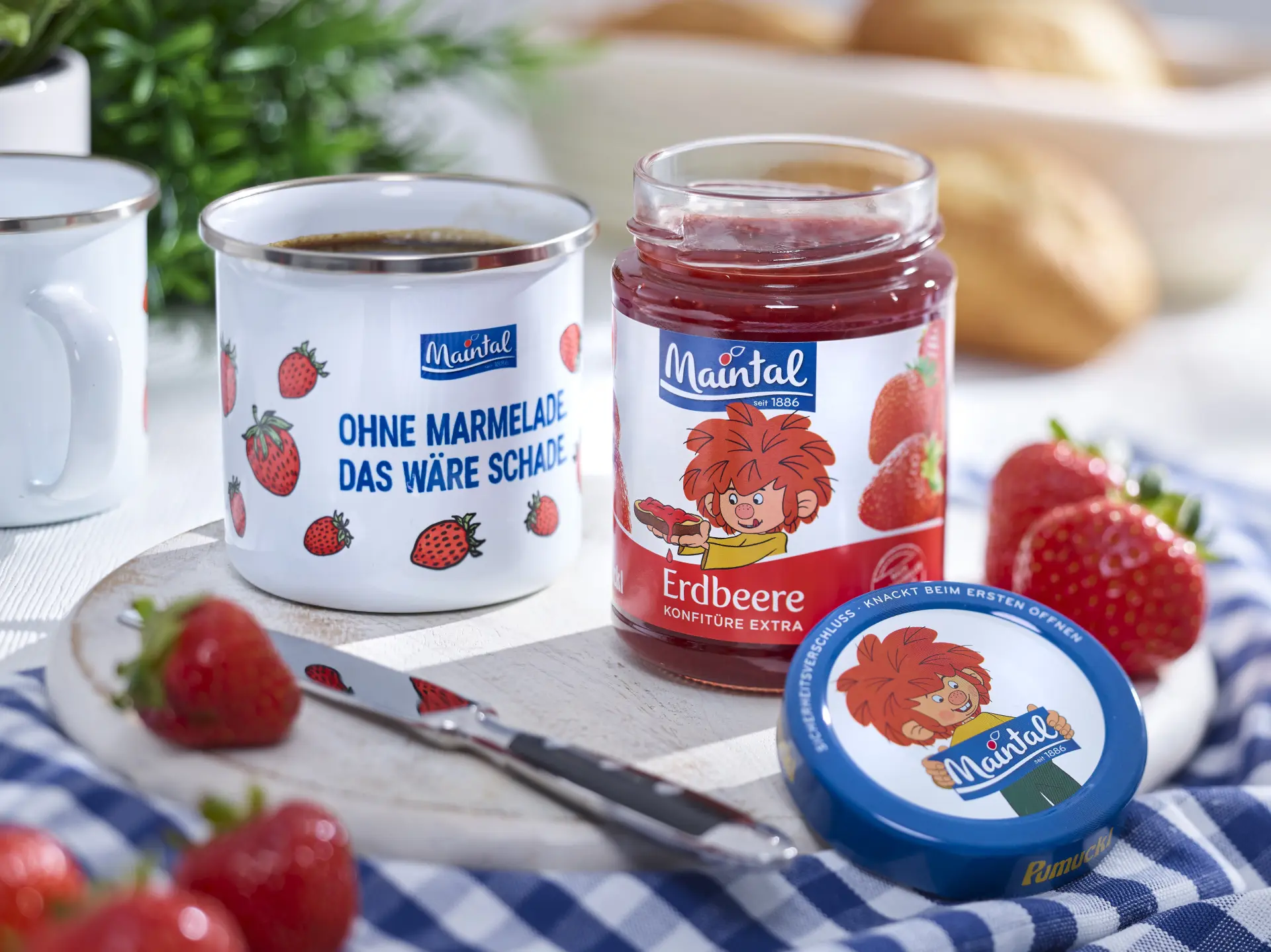 Maintal Pumuckl Erdbeerkonfitüre mit Tasse, Messer und frischen Erdbeeren auf Frühstückstisch.