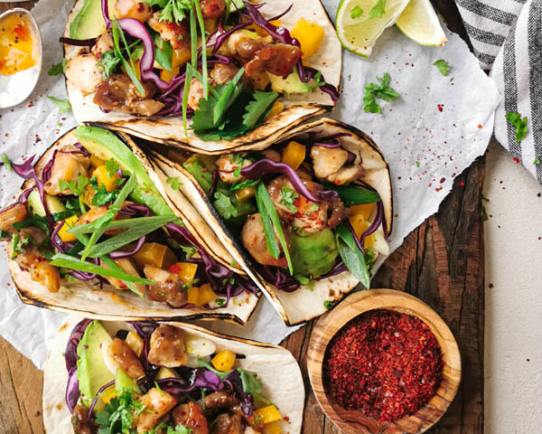 Taccos mit Aprikosen-Haehnchen auf Backpapier und Holzbrett serviert mit Zitronenscheiben und Schüssel mit Gewürz.