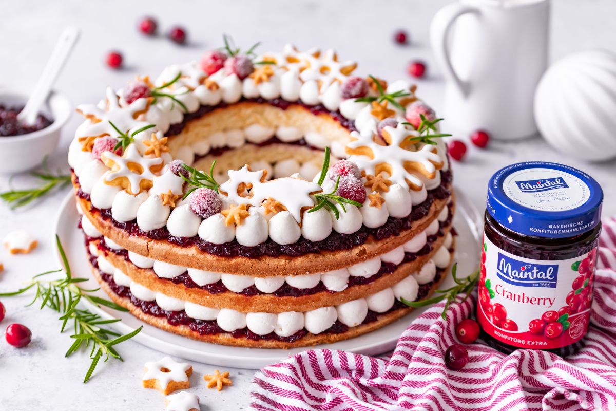 Weihnachtskranz-Torte aus Mandelbiskuit, Cranberries und Vanillecreme Serviervorschlag Weihnachtskranz-Torte auf Tortenplatte, daneben Glas Cranberry-Konfitüre, Milchkännchen, Weihnachtskugel, lose Cranberries.