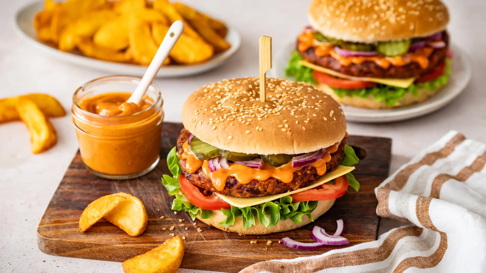 Vegetarischer Burger auf einem Holzbrett mit Kartoffelspalten, einem Glas mit Dip links. Dahinter ein weiterer Burger und eine Schale mit Kartoffelspalten.