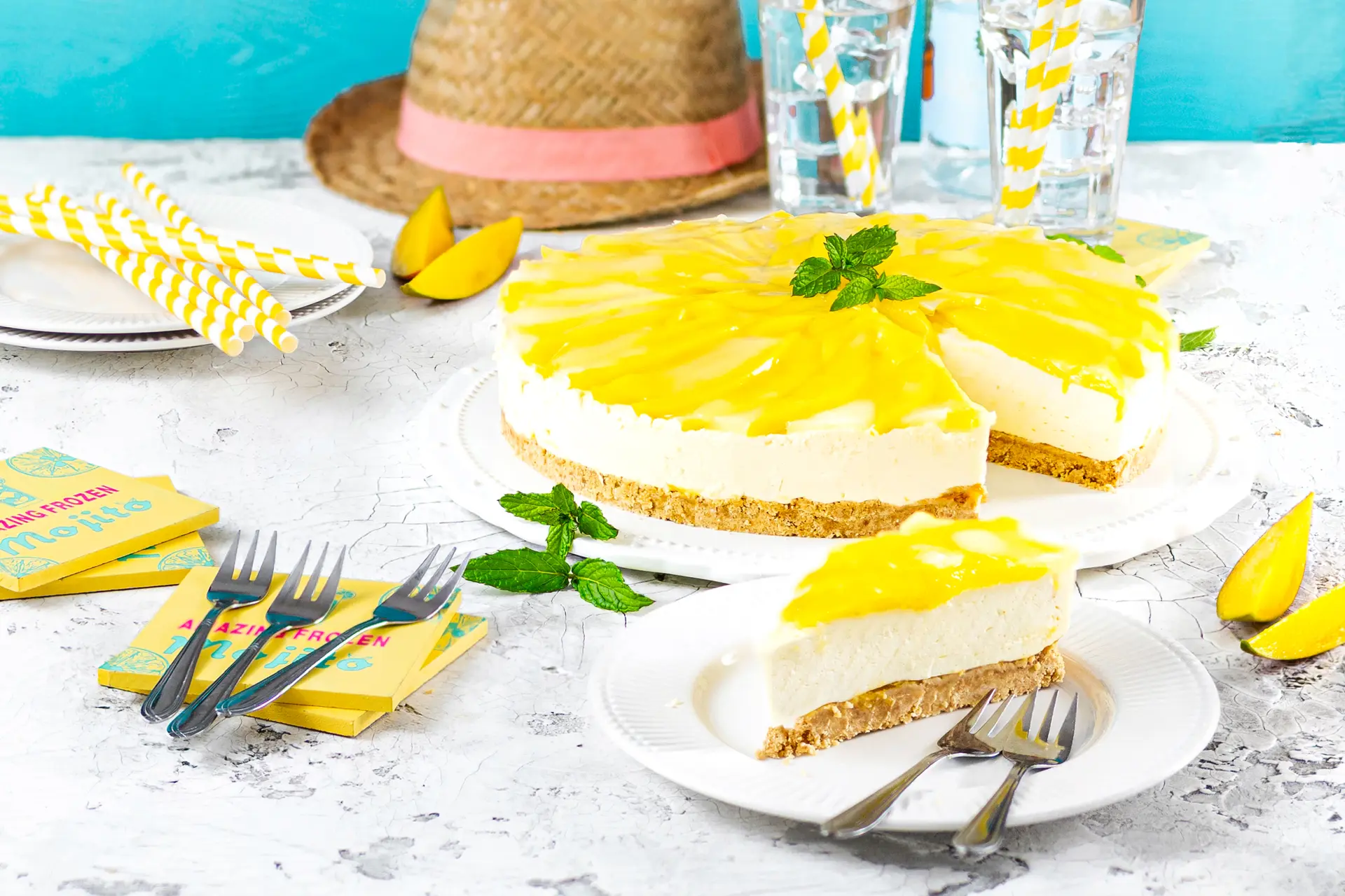 No-Bake-Mango-Cheesecake mit Mangospalten und Minze auf einem Teller, daneben Gabeln, Gläser und Sommerdeko.