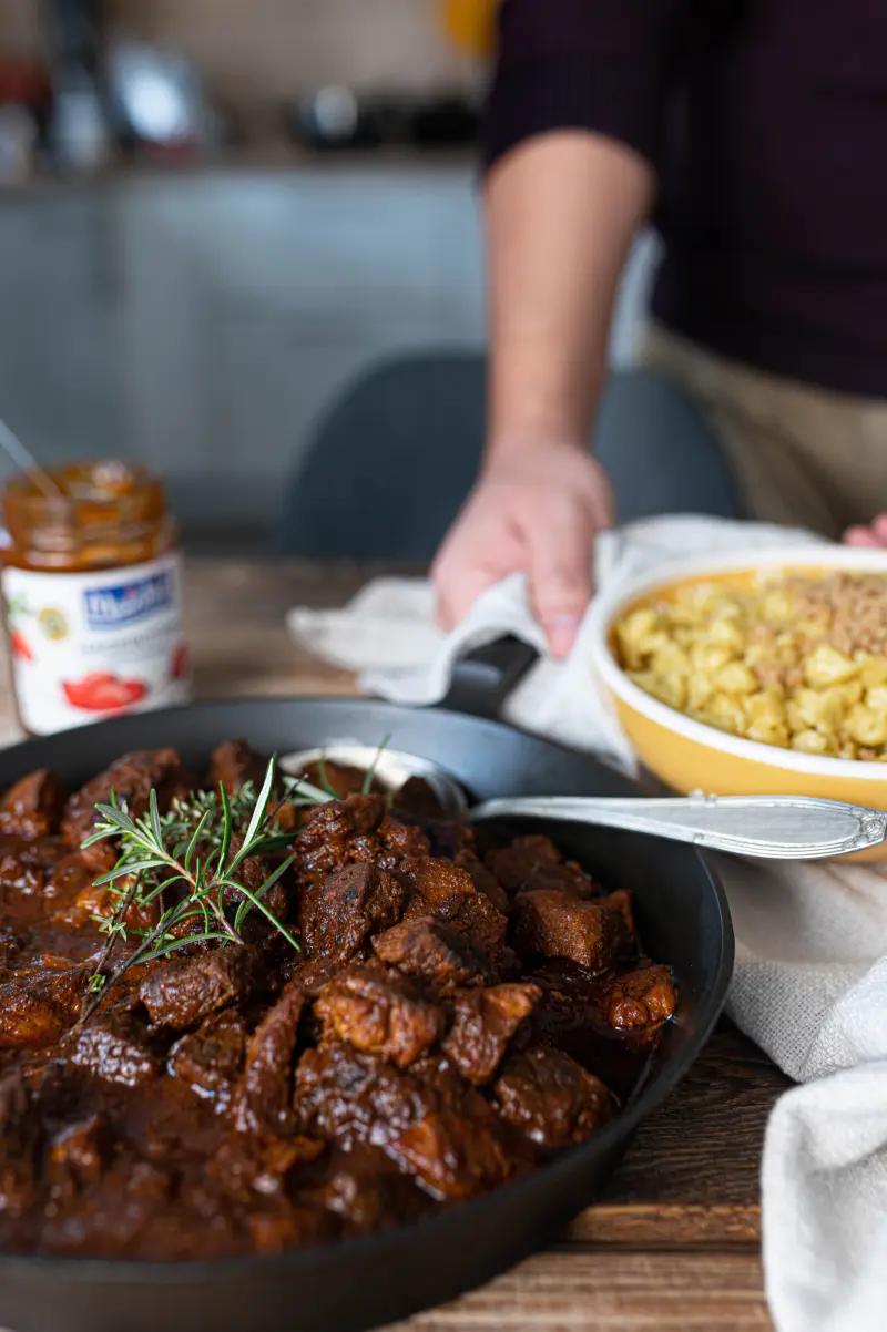 Heiße Pfanne mit Portweingulsch wird mit Handtuch auf Tisch gestellt. Dort sind Glas Hagebutten-Konfitüre und Schüssel mit Spätzle.