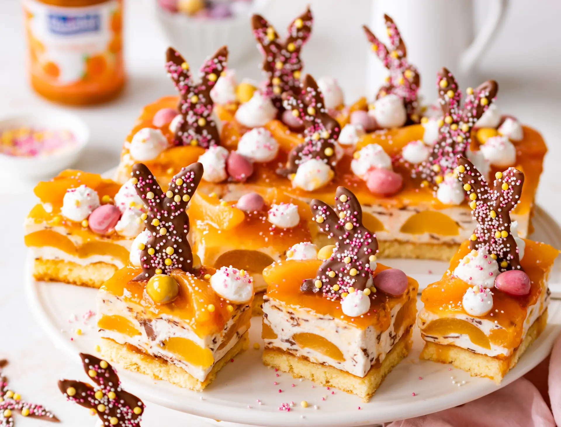 Aprikosen-Blechkuchen mit Stracciatella-Creme – perfekter Osterkuchen Blechkuchen mit Aprikosen und Stracciatella-Creme, dekoriert mit Schokohasen und Streuseln auf einer Tortenplatte.