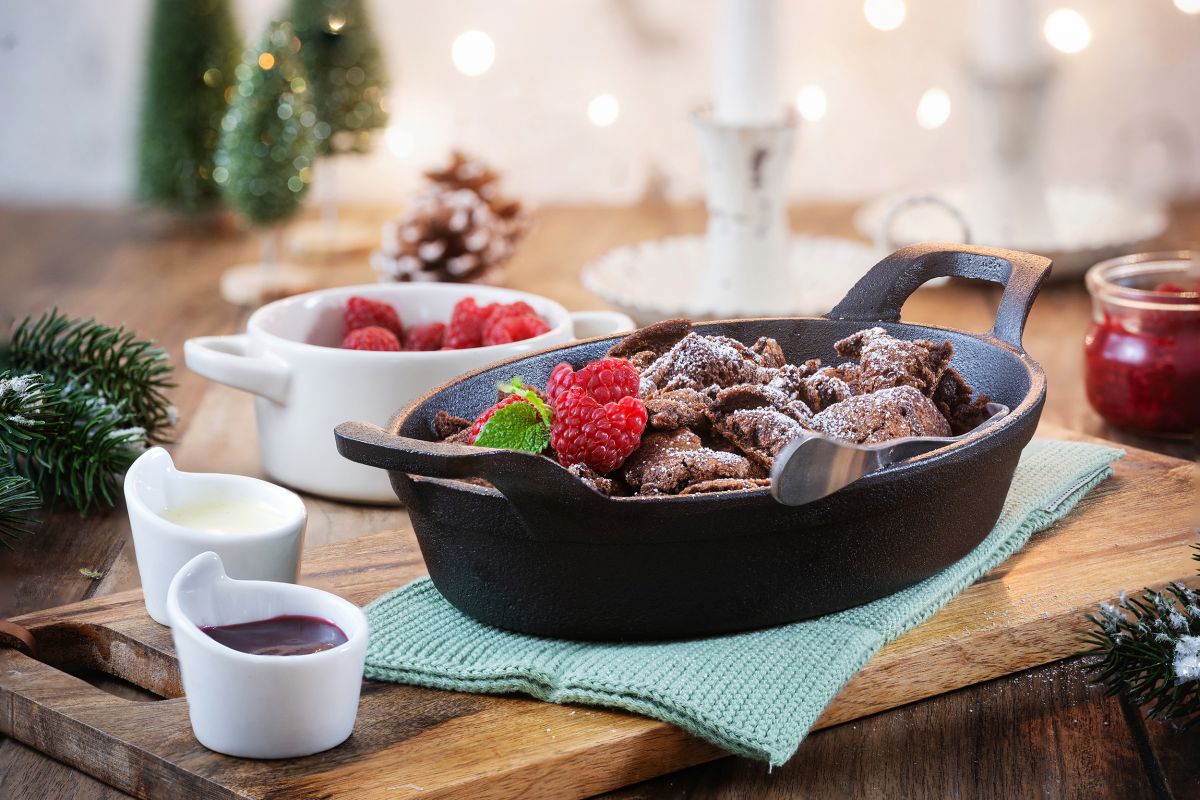 Schokoladen-Kaiserschmarrn mit Himbeeren in Gusseisenpfanne, je ein Schälchen mit Himbeerkompott und Vanillesauce auf weihnachtlich dekoriertem Tisch.