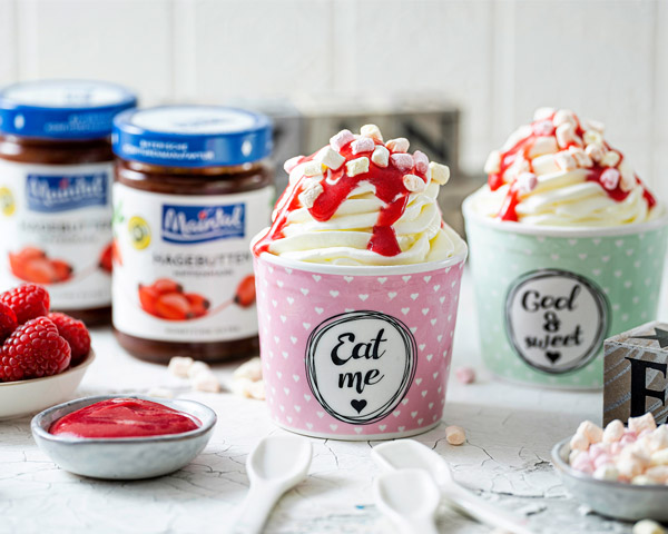 Frozen Joghurt mit Hagebutten-Himbeer-Sauce in Porzellaneisbechern serviert, daneben Schüsselchen mit Sauce, frische Himbeeren, Mini-Marschmallows und Gläser Hagebutten-Konfitüre.