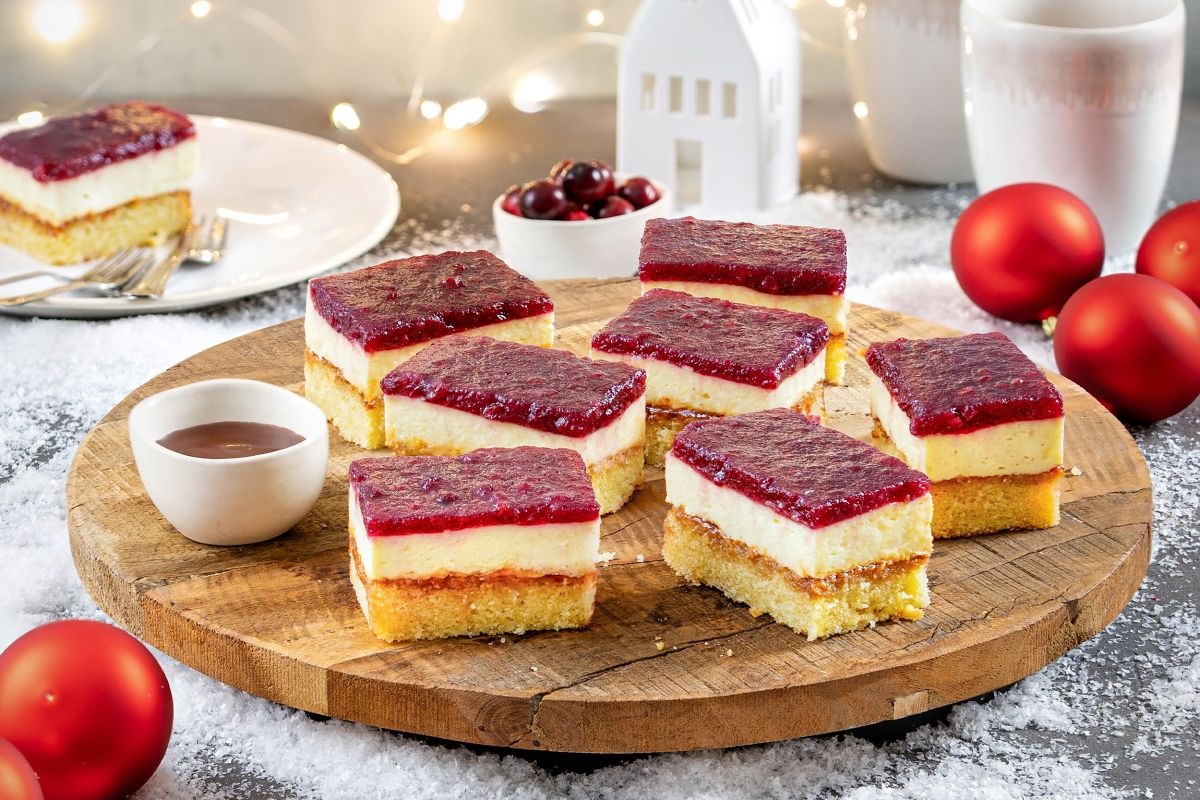 In Stuecke geschnittener Blechkuchen mit Preiselbeer-Gluehweinspiegel auf einer Holzplatte, Teller mit Kuchenstück, Kugeln und winterlicher Deko.