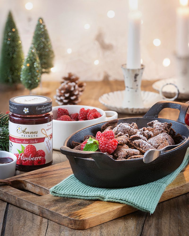 Rezeptbild Schokoladen-Kaiserschmarrn in Gusseisenpfanne, Annes Feinste Bio Himbeeer-Fruchtaufstrich und Schale mit frischen Himbeeren auf weihnachtlich dekoriertem Tisch.