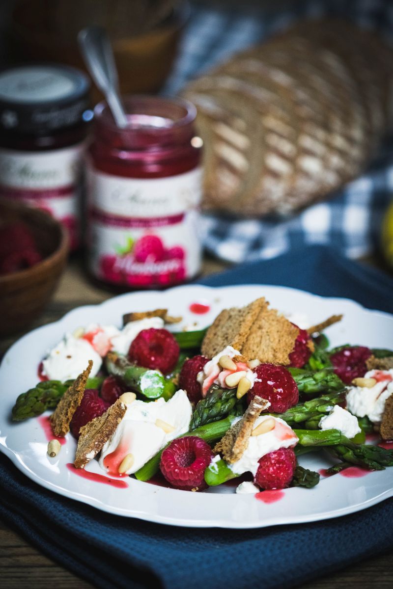 Himbeer-Spargelsalat mit Ricotta und Sauerteigbrotchips Himbeer-Spargel-Salat auf Teller serviert, im Hintergrund Gläser Annes Feinste Bio Himbeer-Fruchtaufstrich und Laib Brot.