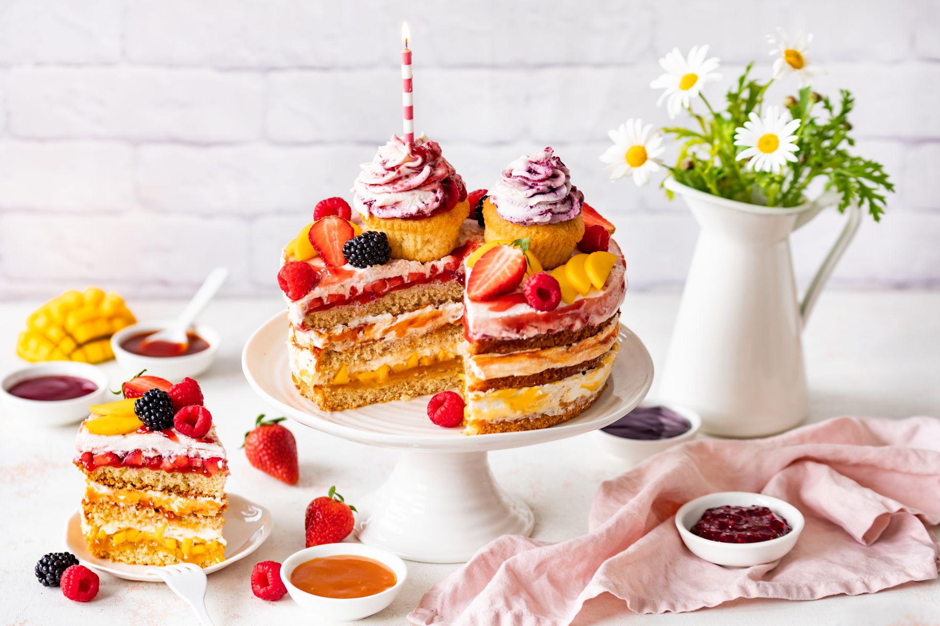 Mehrschichtiger Naked Cake mit Cupcakes, Erdbeeren, Himbeeren, Brombeeren und Mango auf einer weißen Tortenplatte