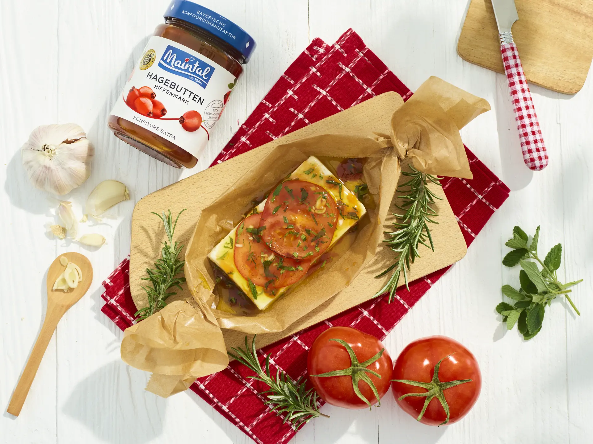 Gegrillter Fetakäse auf Holzbrett in Backpapier mit Tomatenscheiben, Hagebutten-Marinade, Rosmarin und Knoblauch, daneben ein Glas Hagebutten-Konfitüre