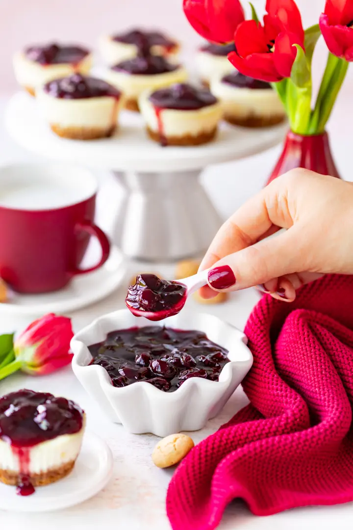 Hand mit rot lackierten Fingernägeln hält einen Löffel mit Sauerkirsch-Konfitüre über eine weiße Schale, im Hintergrund Mini-Cheesecakes mit Kirsch-Topping, eine rote Tasse, Tulpen und ein rotes Tuch.