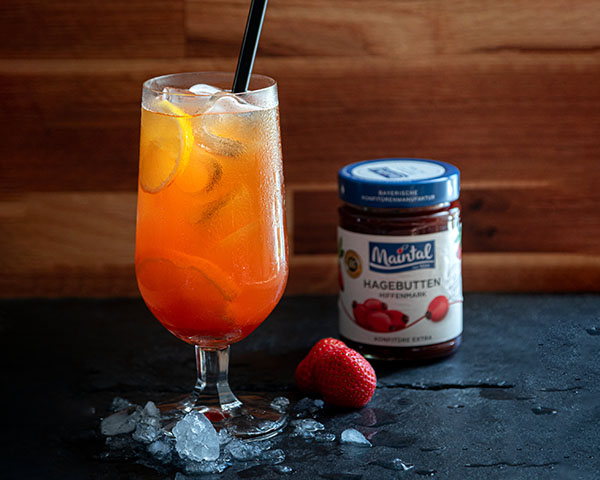 Hagebutten-Shake in hohem Glas mit Strohhalm serviert, daneben ein Glas Hagebutten-Konfitüre und Erdbeeren.