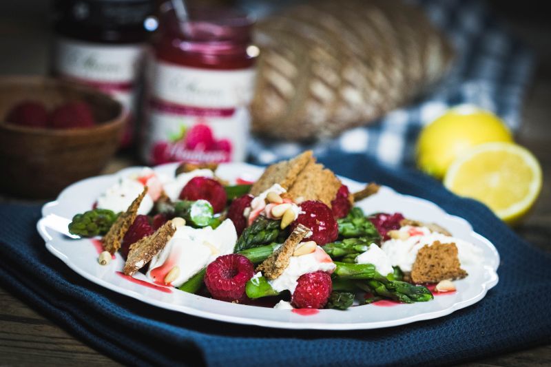 Himbeer-Spargelsalat mit Ricotta und Sauerteigbrotchips Teller mit Himbeer-Spargel-Salat. Im Hintergrund Gläser Annes Feinste Himbeer-Fruchtaufstrich, Zitrone, Laib Brot und Schale mit Himbeeren.