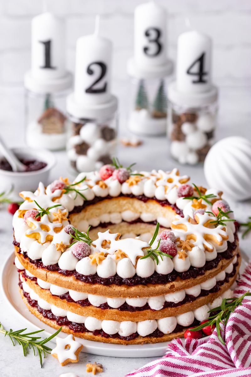 Weihnachtskranz-Torte aus Mandelbiskuit, Cranberries und Vanillecreme Weihnachtskranz-Torte mit Cranberries auf Tortenplatte, daneben Glas Cranberry-Konfitüre, Milchkännchen, Weihnachtskugel, lose Cranberries, vor Adventskranz aus Marmeladengläsern.
