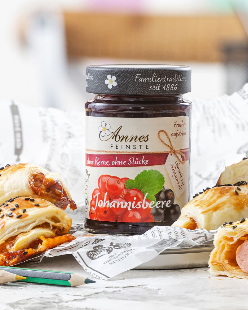 Würstchen im Schlafrock mit Johannisbeeren – BBQ-Sauce Glas Annes Feinste Bio Johannisbeeren-Fruchtaufstrich. Daneben liegen Wuerstchen im Schlafrock.