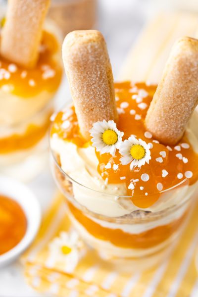 Nahaufnahme Glas mit Schichtdessert mit Eierlikör, Maintal Bio Aprikose-Mango und Granola und Osterhasenohren aus Biskuitstäbchen.