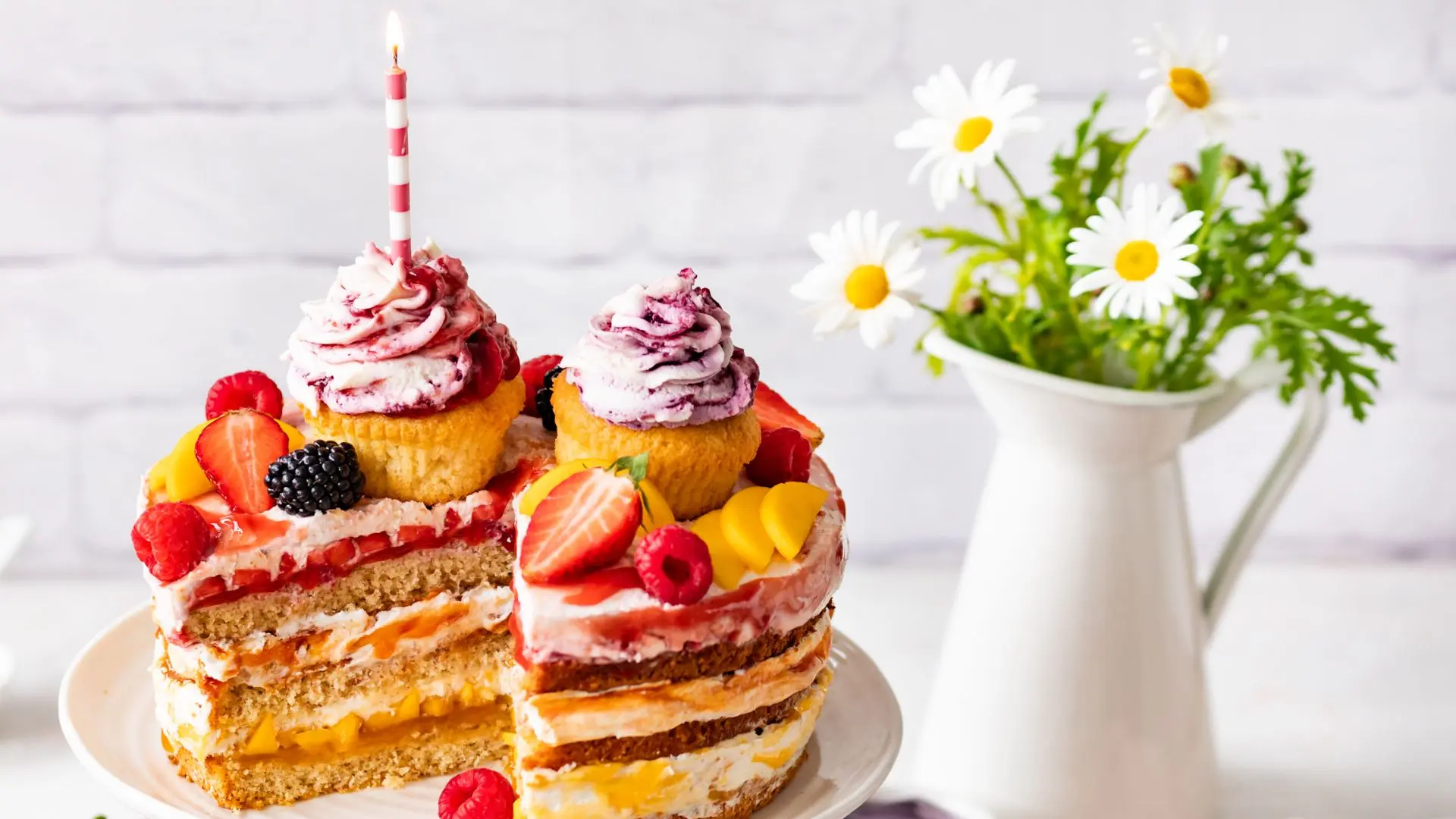 Angeschnittener Naked Cake mit Cupcakes, Beeren, Mango und brennender Kerze