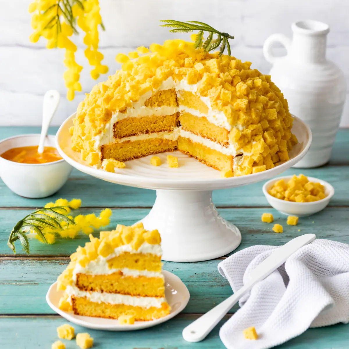 Torta Mimosa mit Zitronencreme und Orangen-Marmelade auf Tortenplatte, daneben ein Strück auf Teller, Schäe mit Orangenmarmelade und eine Schale mit Biskuitwürfeln.