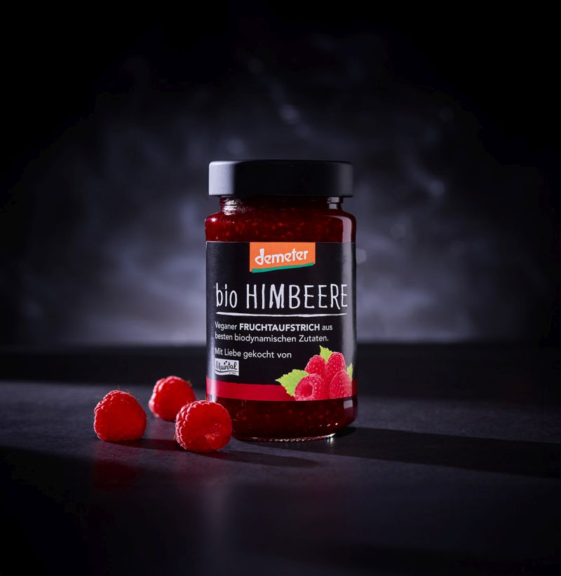 Demeter Bio Fruchtaufstrich Himbeere Glas Maintal Bio Demeter Himbeere-Fruchtaufstrich auf schwarzem Hintergrund, danaben frische Himbeeren.