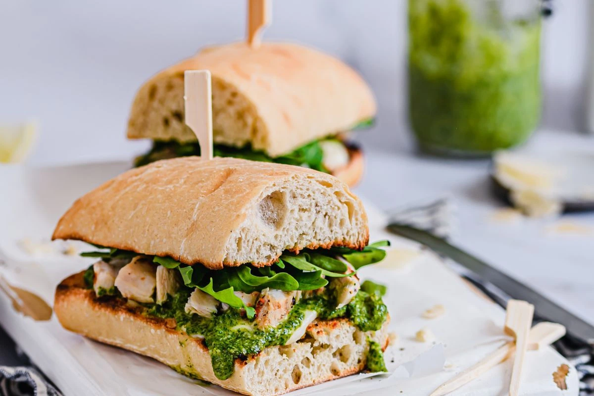 Zwei gegrillte Hähnchen Sandwich mit Pesto auf Platte, daneben Glas mit Pesto.