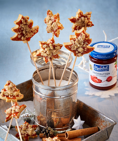 Hagebutten-Streusel-Plätzchen am Stiel Hagebutten-Streusel-Plaetzchen in Blechdose mit Zucker, daneben Glas mit Hagebutten-Konfitüre.