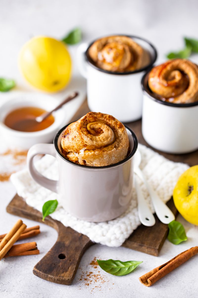 Drei Mug-Cake in der Tasse, auf Brettb serviert, mit Löffeln, Quitten, Zimtstangen und Schüssel mit Quittengelee mit Löffel.